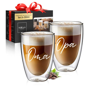 Zwei doppelwandige ElbFuchs Gläser mit den Gravuren 'Oma' und 'Opa', gefüllt mit Latte macchiato, stehen vor einer eleganten schwarzen Geschenkbox mit roter Schleife.