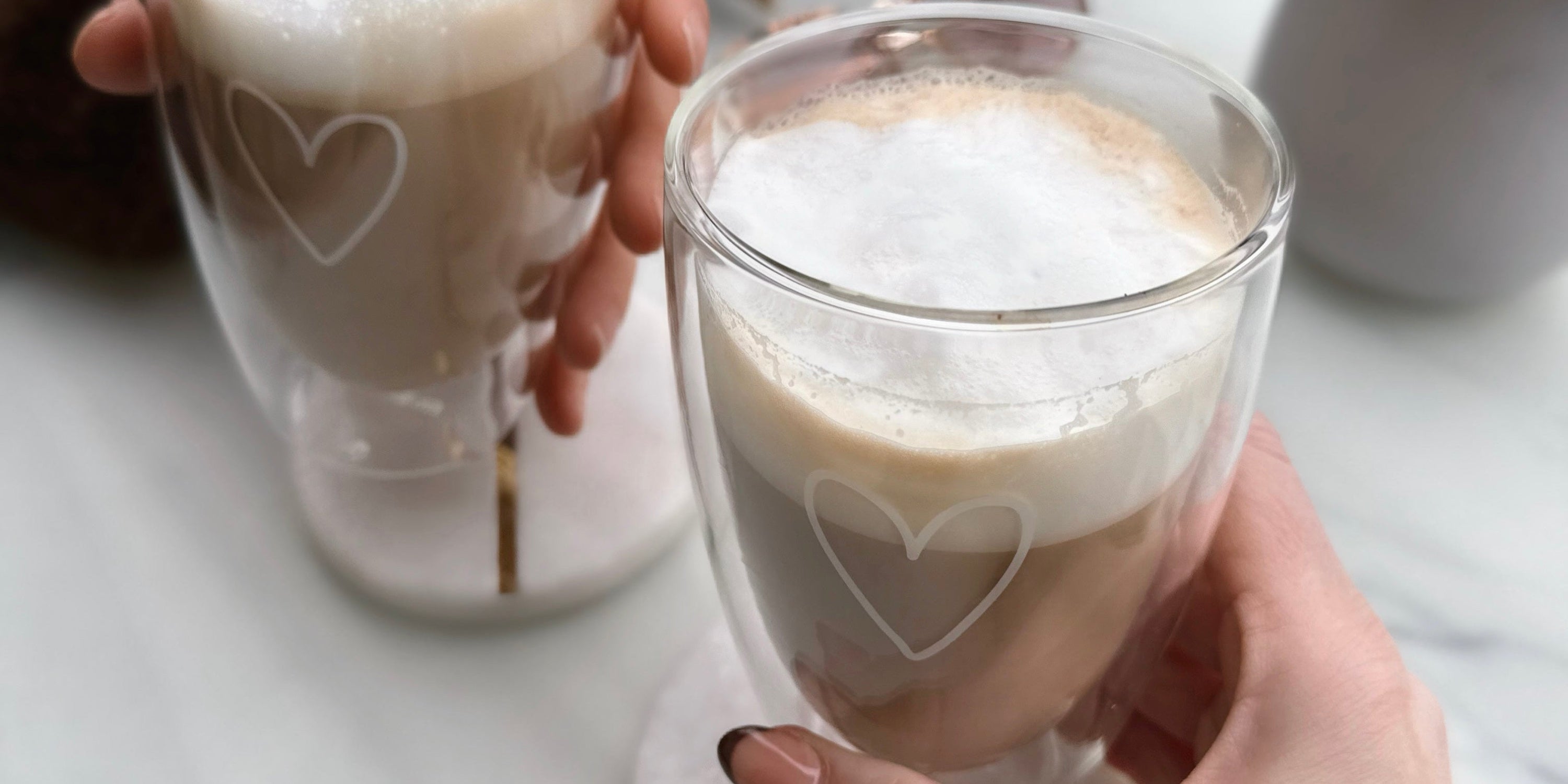 Nahaufnahme von zwei doppelwandigen ElbFuchs Cappuccinogläsern aus der Kollektion 'PARIS', die jeweils eine feine, weiße Herzgravur auf der Außenseite tragen. Zwei Hände halten die Gläser, welche mit frisch zubereitetem Cappuccino und einer dichten, cremigen Schicht aus weißem Milchschaum befüllt sind. Der helle Hintergrund in dezenter Marmoroptik unterstreicht das moderne, ästhetische Design dieses Lifestyle-Produktfotos für den Kaffeegenuss zu Hause.