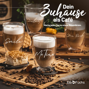 Vier doppelwandige ElbFuchs Gläser mit Latte Macchiato stehen auf einer Bambusmatte zwischen Kaffeebohnen und Gebäck, in der Mitte mit der Gravur 'Me Time' und im Hintergrund angedeutet die Schriftzüge 'Soulmate' und 'You Rock'.