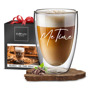 Ein ElbFuchs Doppelwandiges Glas 350ml mit der Gravur 'Me Time', gefüllt mit cremigem Latte Macchiato, steht vor einer schwarzen Geschenkbox mit roter Schleife und dem ElbFuchs Logo.