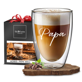 Ein ElbFuchs Doppelwandiges Glas 350ml mit der Gravur 'Papa', gefüllt mit cremigem Latte Macchiato, steht vor einer schwarzen Geschenkbox mit roter Schleife und ElbFuchs Logo.