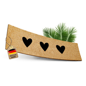 ElbFuchs Kokos-Fußmatte 75x25 cm mit drei schwarzen Herz-Cutouts, leicht gebogen freigestellt vor weißem Hintergrund, mit Anhänger 'Deutsche Marke' und dekorativen Palmblättern.