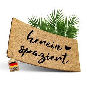 Im hellen Flur liegt die ElbFuchs Kokosfußmatte „herein spaziert“ mit Herz vor einer schmaleren Schuhmatte mit Sneakern, neben einer Vase mit Trockenblumen und einem gedeckten Esstisch im Hintergrund.