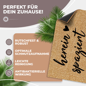 Eine hochgeklappte ElbFuchs Kokosfußmatte mit dem Schriftzug „herein spaziert“ und Herz zeigt ihre schwarze rutschfeste Unterseite vor Palmblättern, dazu Hinweise zu Robustheit, Schmutzaufnahme, leichter Reinigung und antibakterieller Wirkung.