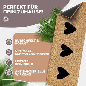Nahaufnahme einer hochgeklappten ElbFuchs Kokos-Fußmatte mit drei Herz-Ausschnitten, die die schwarze rutschfeste Gummiunterseite zeigt, dekoriert mit Palmblättern. Symbole daneben verweisen auf Rutschfestigkeit, hohe Schmutzaufnahme, leichte Reinigung und antibakterielle Wirkung.