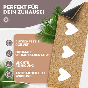 Eine ElbFuchs Kokos-Fußmatte mit weißen Herzen ist teilweise aufgerollt, sodass die rutschfeste schwarze Gummirückseite sichtbar wird; Palmblätter im Hintergrund unterstreichen die natürliche Optik.