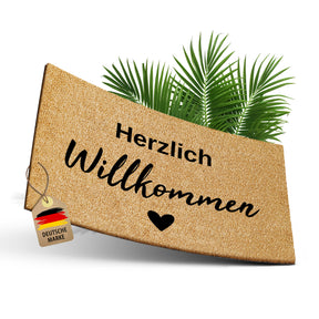 Freigestellte ElbFuchs Kokosfußmatte mit der Gravur „Herzlich Willkommen“ und Herz vor grünen Palmblättern, mit einem Anhänger „Deutsche Marke“ als Hinweis auf die heimische Fertigung.