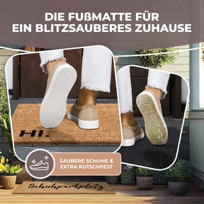 Nahaufnahme zweier Schuhsohlen beim Abstreifen auf der ElbFuchs Kokos-Fußmatte mit der Aufschrift 'HI.', mit dem Hinweis 'Saubere Schuhe & extra rutschfest' vor einem unscharfen Hauseingang.