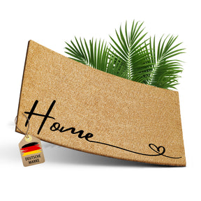 Freigestellte ElbFuchs Kokosfußmatte mit dem Schriftzug „Home“ und einem herzförmigen Schwung am Ende vor Palmblättern, mit Anhänger „Deutsche Marke“.