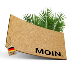 Freigestellte ElbFuchs Kokos-Fußmatte 60x40 cm mit der Aufschrift 'MOIN.' in schwarzer Schrift, dekoriert mit grünen Palmblättern im Hintergrund.