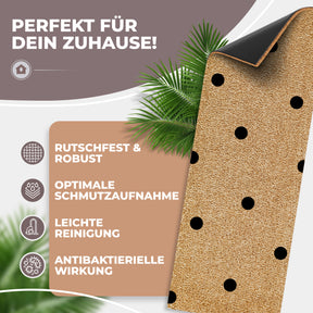 Eine ElbFuchs Kokos-Fußmatte mit schwarzen Punkten ist teilweise aufgerollt, sodass die rutschfeste schwarze Gummirückseite sichtbar wird; Palmblätter im Hintergrund unterstreichen die natürliche Optik.