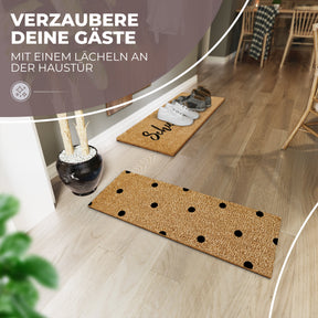 In einem hellen Eingangsbereich mit Holzdielenboden liegt eine ElbFuchs Kokosmatte mit schwarzen Punkten neben einer schwarzen Vase mit Trockenzweigen; im Hintergrund steht die passende Schuhparkplatz-Matte mit ordentlich aufgereihten Schuhen vor einem gedeckten Esstisch.