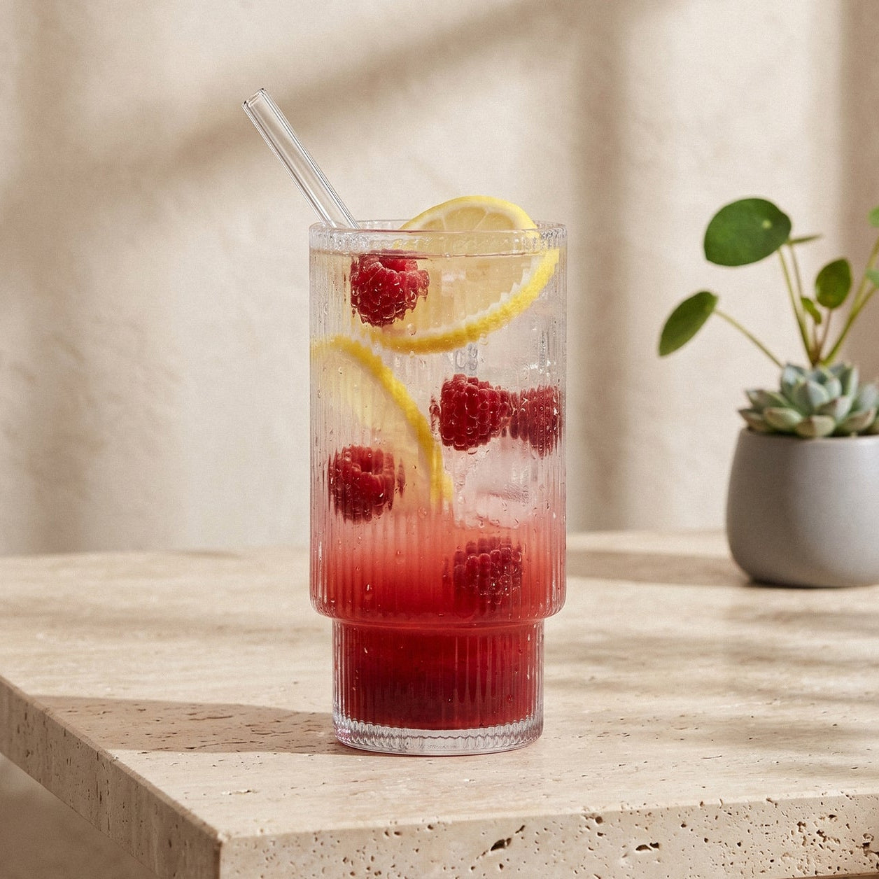 Das ElbFuchs RILA Highball-Glas präsentiert sich als stilvolles, geriffeltes Trinkglas mit charakteristischer vertikaler Rillen-Textur im unteren Bereich und einem glatten oberen Rand. Befüllt mit einem erfrischenden Frucht-Drink aus frischen Himbeeren, Zitronenscheiben, Eiswürfeln und einem gebogenen Glas-Strohhalm, steht das elegante Longdrinkglas mittig auf einem hellen Travertin-Steintisch. Natürliches Sonnenlicht wirft sanfte Schatten auf die sandfarbene Wand im Hintergrund.