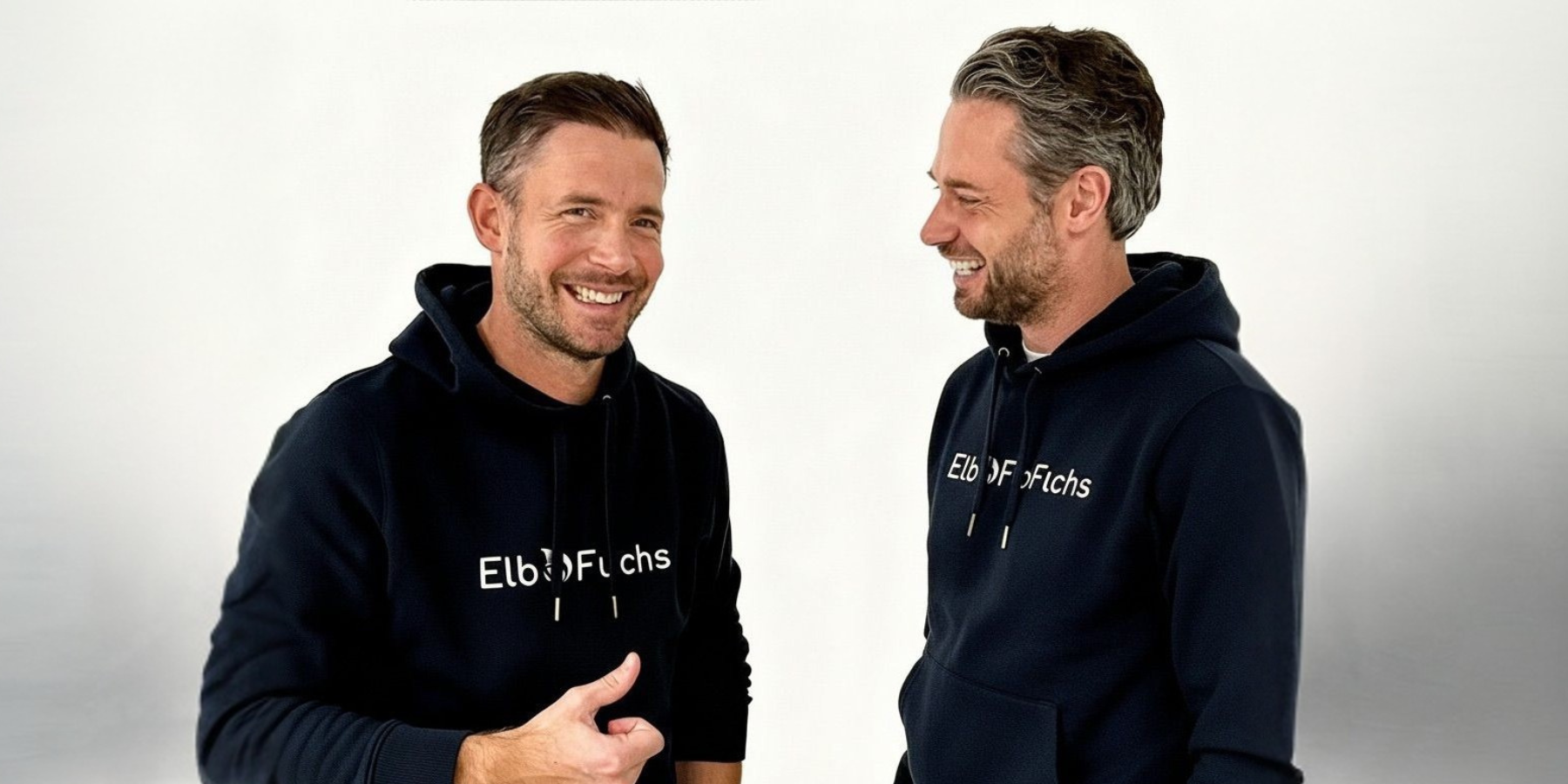 Authentisches Gründerfoto der ElbFuchs-Gründer Christoph und Kevin aus Hamburg. Die beiden Männer blicken sich an und lachen herzlich vor einem cleanen, weißen Studiohintergrund. Sie tragen farblich passende, dunkelblaue Hoodies mit dem weißen ElbFuchs-Markenlogo auf der Brust.