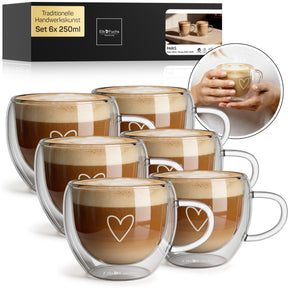Sechs ElbFuchs PARIS Cappuccino-Tassen mit doppelwandigem Glas, Henkel und feiner Herzgravur, gefüllt mit cremigem Milchkaffee im Set 6x 250ml, mit Verpackung und einer Nahaufnahme haltender Hände im Hintergrund.