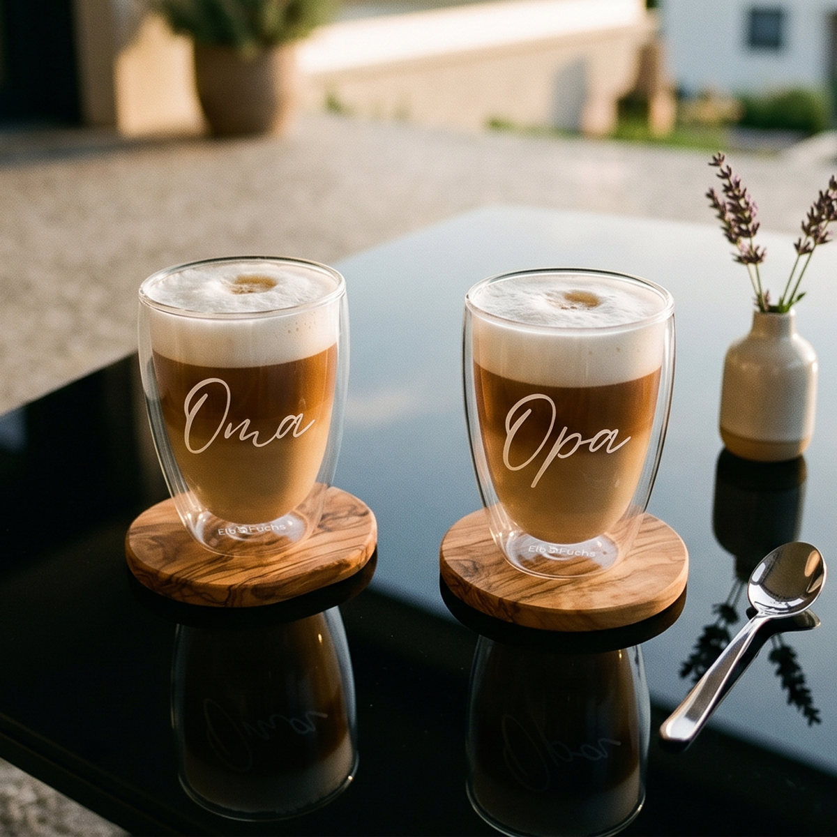 Zwei ElbFuchs doppelwandige Kaffeegläser (350ml) aus der WORDS Kollektion mit feiner weißer Gravur der Schriftzüge 'Oma' und 'Opa'. Die mit geschichtetem Latte Macchiato und dichtem Milchschaum befüllten Thermogläser stehen auf runden Olivenholz-Untersetzern auf einem dunkel spiegelnden Tisch im Freien. Eine liebevolle Geschenkidee für werdende Großeltern.