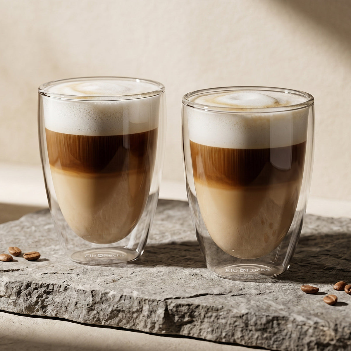 ElbFuchs VIENNA doppelwandiges Kaffeeglas-Set (350ml) aus Borosilikatglas, gefüllt mit geschichtetem Latte Macchiato und einer dichten, cremigen Milchschaumhaube. Die zwei Thermo-Kaffeegläser stehen auf einer rustikalen, grauen Natursteinplatte mit verstreuten Kaffeebohnen im warmen Streiflicht. Isolierende Gläser für ein stilvolles Kaffeeritual.