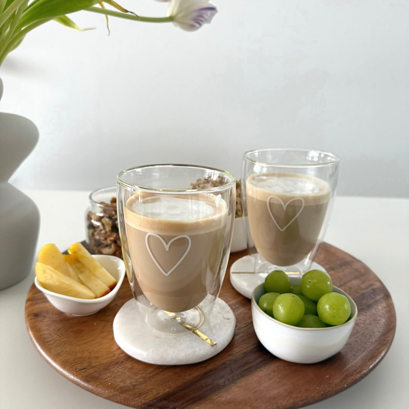 Zwei doppelwandige ElbFuchs Kaffeegläser aus der PARIS Kollektion mit einer feinen weißen Herzgravur, befüllt mit cremigem Milchkaffee. Die Thermogläser stehen auf runden, weißen Marmoruntersetzern mit Goldakzent auf einem runden Serviertablett aus Holz. Dekoriert ist die gemütliche Frühstücksszene mit Schälchen voller grüner Weintrauben, Apfelspalten und Nüssen vor einem hellen Hintergrund mit einer Blumenvase.