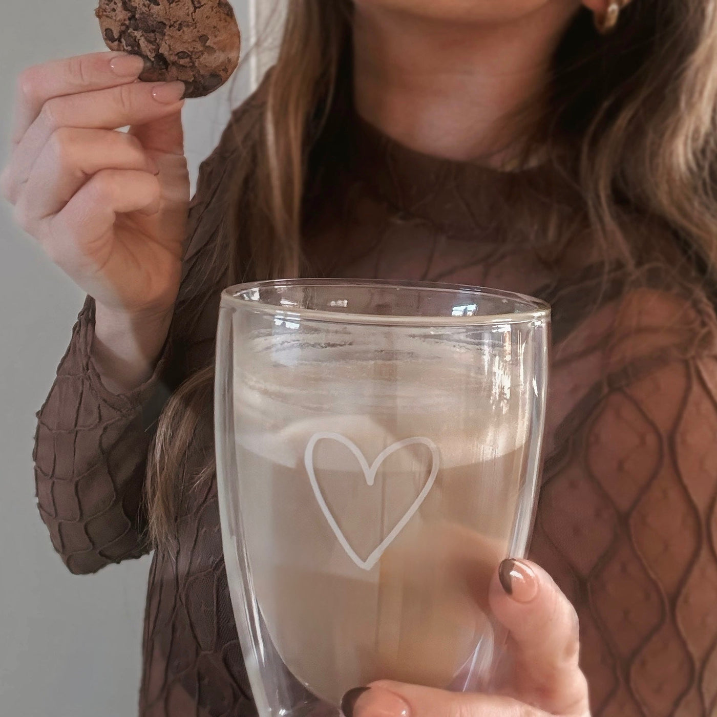 Ein ästhetisches Lifestyle-Foto im Nahbereich: Eine Frau in einem braunen, transparent gemusterten Oberteil hält ein doppelwandiges ElbFuchs Kaffeeglas aus der PARIS Kollektion mit einer feinen, weißen Herzgravur. Das Thermoglas ist mit cremigem Milchkaffee befüllt, während sie in der anderen Hand einen Schokoladen-Cookie hält. 