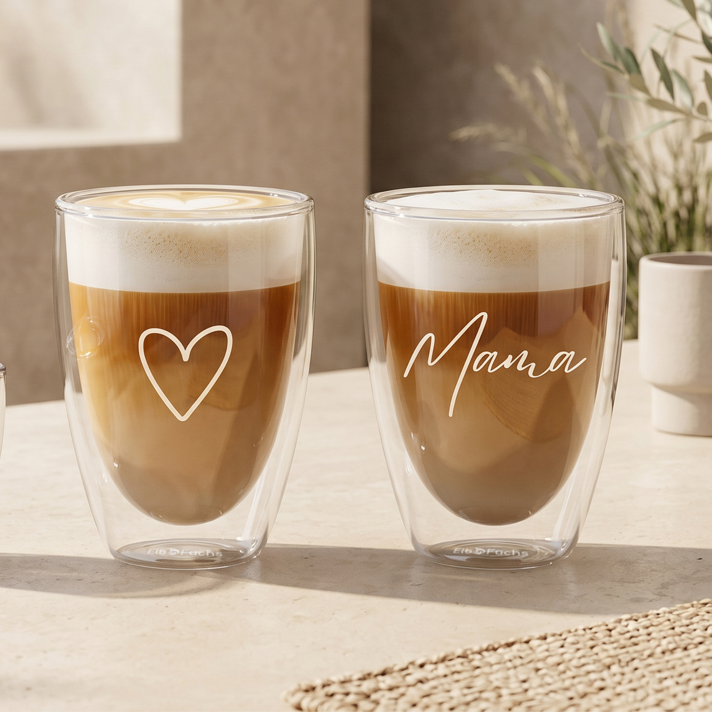 Zwei ElbFuchs doppelwandige Latte-Macchiato-Gläser (350ml) mit feiner weißer Gravur: Ein Thermoglas mit einem Herz-Symbol und ein Glas mit dem eleganten Schriftzug 'Mama'. Die mit geschichtetem Kaffee und dichtem Milchschaum befüllten Gläser stehen auf einem hellen Tisch im warmen, natürlichen Sonnenlicht. Ein perfektes Geschenkset zum Muttertag.