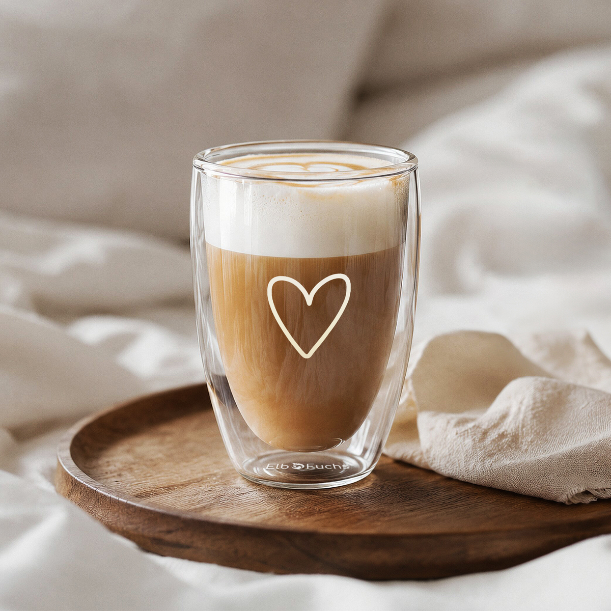 Ein ElbFuchs doppelwandiges Kaffeeglas (350ml) der Kollektion PARIS mit feiner weißer Herzgravur, befüllt mit einem cremigen Cappuccino und sanfter Latte-Art auf dem Milchschaum. Das Thermo-Kaffeeglas steht auf einem runden Holztablett neben einem hellen Leinentuch inmitten eines gemütlich gemachten Betts mit weißen Decken.
