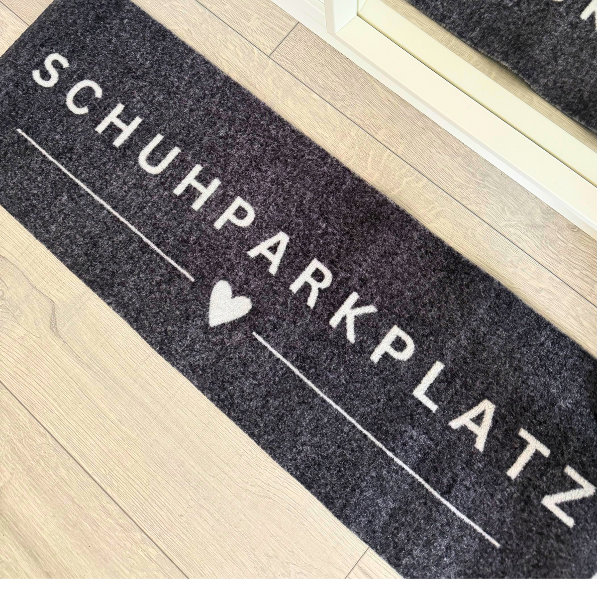 Eine dunkelgraue ElbFuchs Filz-Schuhmatte mit dem markanten, weißen Schriftzug 'SCHUHPARKPLATZ' und einem integrierten Herz-Symbol. Die stilvolle Schuhablage liegt auf einem hellen Fußboden in Holzoptik im Flur oder Eingangsbereich und dient als sauberer Abstellplatz für Schuhe.