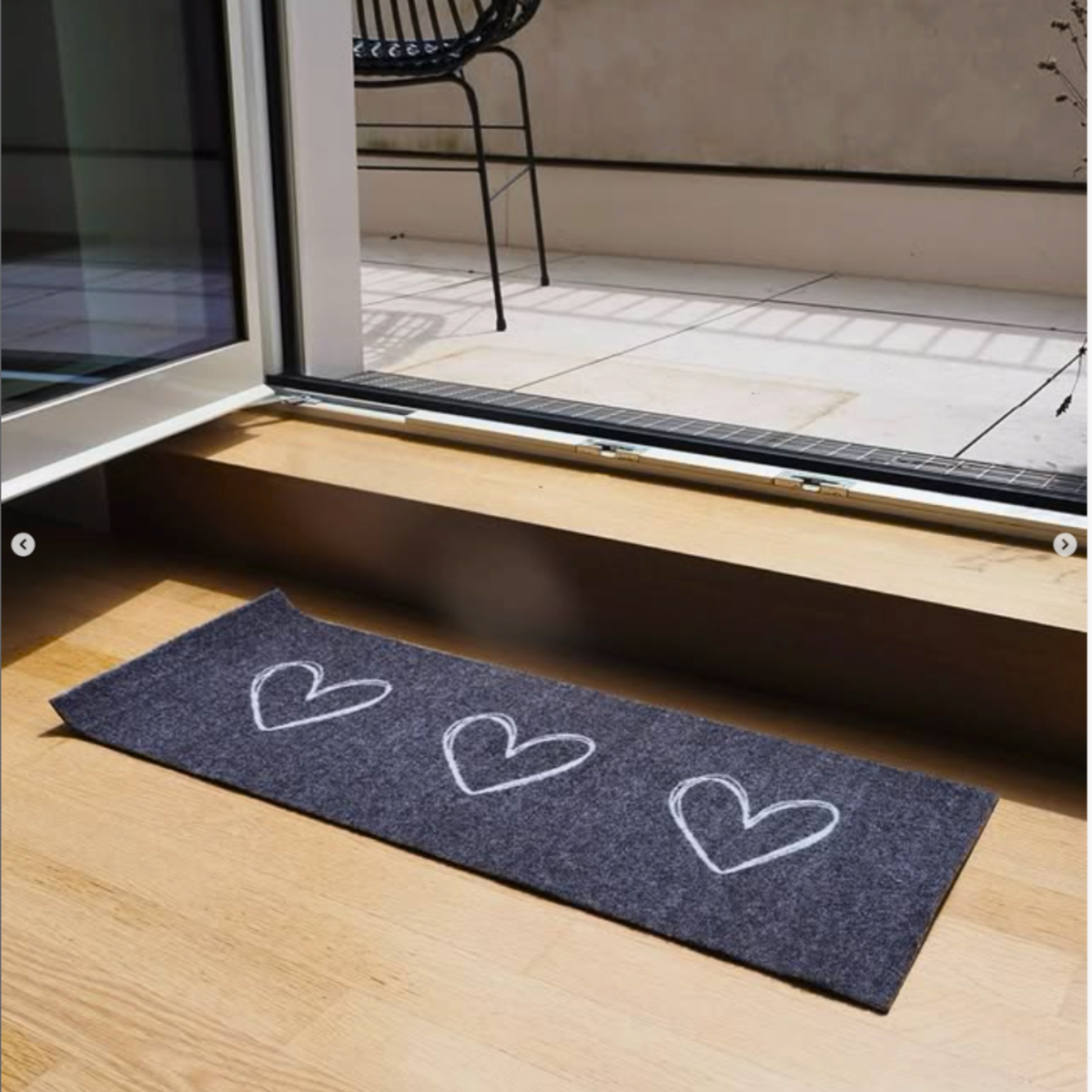 Eine schmale, dunkelgraue ElbFuchs Velour-Schuhmatte mit drei minimalistisch gezeichneten, weißen Herzen. Die elegante Schuhunterlage liegt auf einem hellen Holzboden direkt vor einer geöffneten Terrassentür oder Balkontür und dient als stilvolle Schmutzfangmatte und dekorative Schuhablage für den Innenbereich. Hergestellt in Deutschland aus recycelten Kunststoffen.