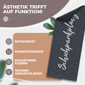 Eine aufgerollte anthrazitfarbene ElbFuchs Schuhparkplatz Fußmatte mit elegantem Schreibschrift-Schriftzug und sichtbarer rutschfester Unterseite, dekoriert mit grünen Blättern; Icons zeigen Rutschfestigkeit, Schmutzfang und Wasserundurchlässigkeit.