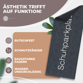 Eine aufgerollte anthrazitfarbene ElbFuchs Schuhparkplatz Fußmatte mit sichtbarer rutschfester schwarzer Unterseite, dekoriert mit grünen Blättern; Icons weisen auf Rutschfestigkeit, Schmutzfang und Wasserundurchlässigkeit hin.