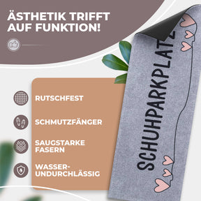 Eine aufgerollte graue ElbFuchs Schuhparkplatz Fußmatte mit vertikalem Schriftzug und rosa Herzen sowie sichtbarer schwarzer, rutschfester Unterseite vor grünen Blättern; Icons zeigen Rutschfestigkeit, Schmutzfang und Wasserundurchlässigkeit.