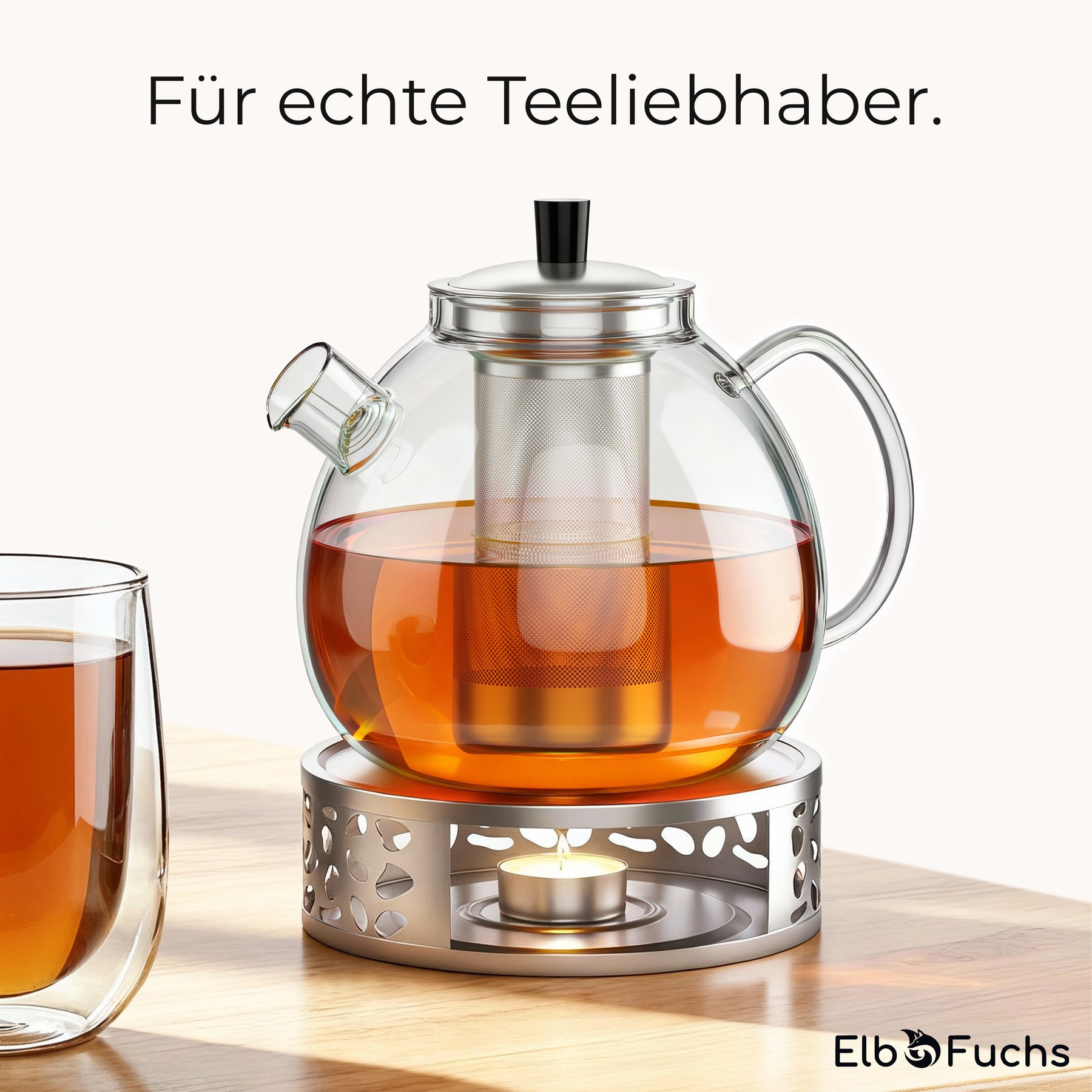 Die ElbFuchs Glasteekanne mit Edelstahl-Siebeinsatz steht gefüllt mit goldbraunem Tee auf ihrem durchbrochenen Stövchen mit brennendem Teelicht, daneben ein bereits eingeschenktes doppelwandiges Glas.