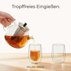 Eine Hand gießt aus der ElbFuchs Glasteekanne mit Edelstahlsieb tropffrei Tee in ein doppelwandiges Glas, während ein zweites, leeres Glas daneben auf dem Holztisch steht.