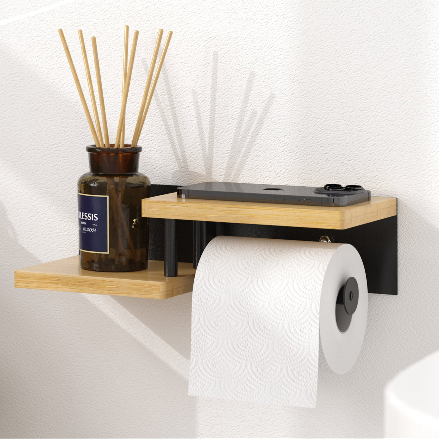 Ein ElbFuchs Toilettenpapierhalter 'Singapor' aus hellem Bambusholz und mattschwarzem Stahl, montiert an einer glatten, weißen Badezimmerwand im warmen Sonnenlicht. Die untere, versetzte Holzablage ist mit einem braunen Flakon samt Duftstäbchen (Raumdiffuser) dekoriert, während auf der oberen Ablage ein schwarzes Smartphone liegt. Links im Bild ist der weiße Spülkasten einer Toilette zu erkennen, auf dem ordentlich gestapelte, weiße Gästehandtücher liegen.