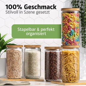 Fünf ElbFuchs Vorratsgläser mit Bambusdeckel stehen gestapelt vor einer weißen Fliesenwand, gefüllt mit Haferflocken, Reis, Kaffeebohnen, Nudeln und bunten Frühstücksringen, dekoriert mit einer grünen Rankpflanze.