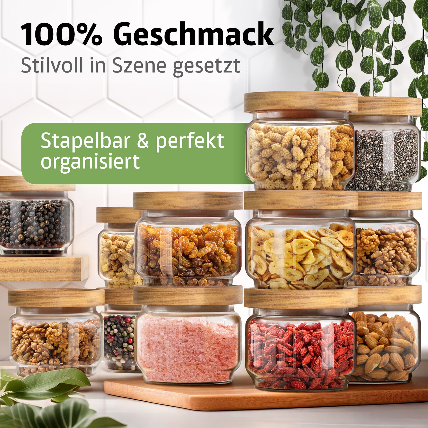 Zahlreiche ElbFuchs Vorratsgläser WOOD KONTOR mit Holzdeckeln sind pyramidenförmig gestapelt und mit Nudeln, Chiasamen, Pfefferkörnern, Rosinen, Bananenchips, Walnüssen, rosa Salz, Gojibeeren und Mandeln gefüllt, vor einer weißen Fliesenwand mit hängenden Ranken.