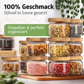 Zahlreiche ElbFuchs Vorratsgläser WOOD KONTOR mit Holzdeckeln sind pyramidenförmig gestapelt und mit Nudeln, Chiasamen, Pfefferkörnern, Rosinen, Bananenchips, Walnüssen, rosa Salz, Gojibeeren und Mandeln gefüllt, vor einer weißen Fliesenwand mit hängenden Ranken.