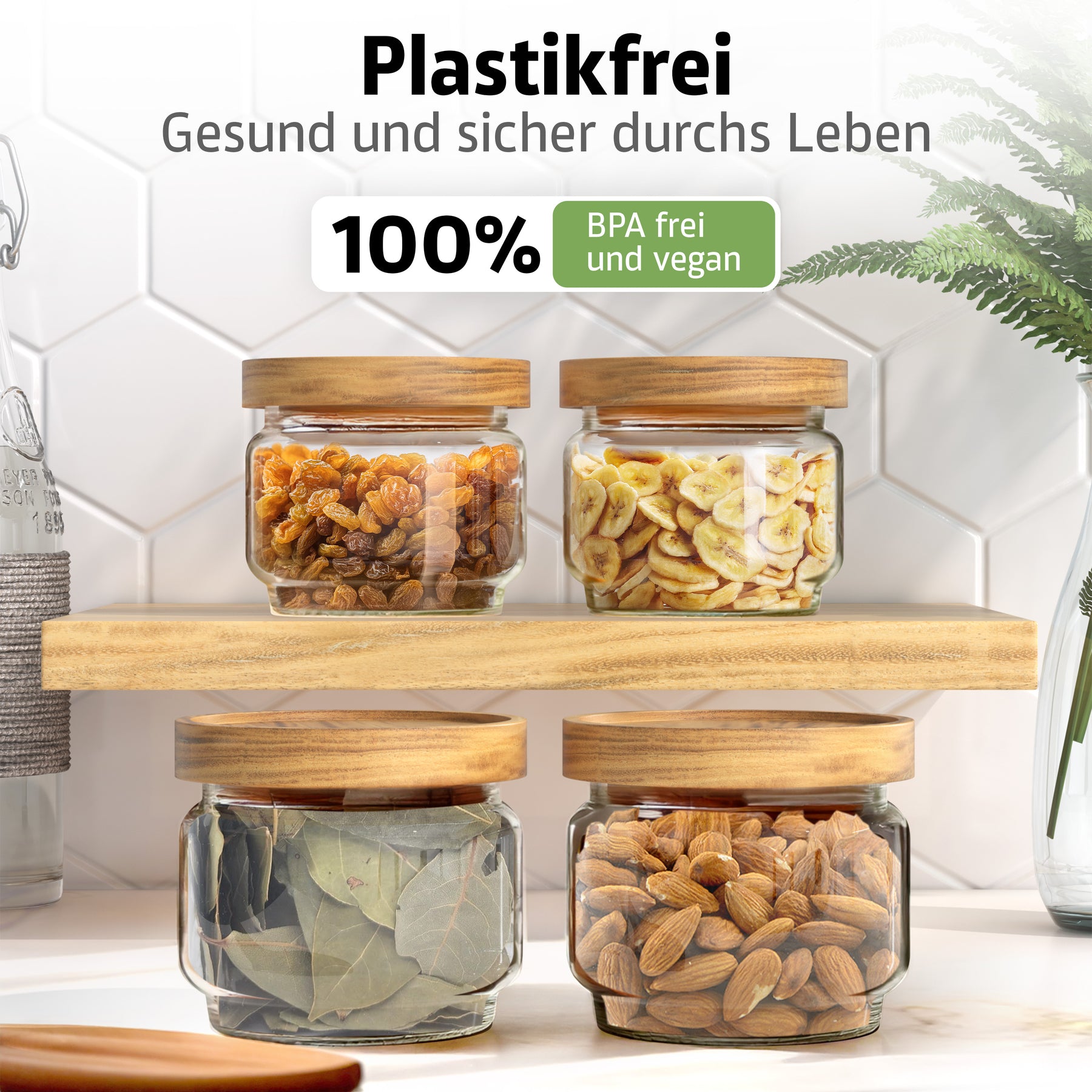 Vier ElbFuchs Vorratsgläser WOOD KONTOR mit Holzdeckeln stehen auf einem Holzbrett, gefüllt mit Rosinen, Bananenchips, Lorbeerblättern und Mandeln, vor weißen Hexagonfliesen mit grüner Pflanze.