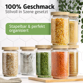 Sieben ElbFuchs Vorratsgläser WOOD KONTOR mit Holzdeckeln sind pyramidenförmig gestapelt und mit Mais, Hirse, Popcornmais, Mehl, grünen Erbsen, roten Linsen und braunem Zucker gefüllt, vor einer weißen Fliesenwand mit hängenden Ranken.