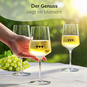 Eine Hand greift nach einem von drei ElbFuchs Weingläsern mit Herzgravur, die mit kühlem Weißwein gefüllt auf einem gedeckten Tisch neben grünen Trauben stehen, im Hintergrund ein sonniger, grün schimmernder Garten.