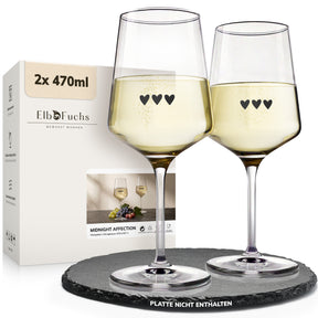 Zwei ElbFuchs Weingläser 'Midnight Affection' mit 470 ml Fassungsvermögen und drei kleinen Herzgravuren stehen gefüllt mit Weißwein neben der Verpackung des 2er-Sets auf einer runden dunklen Schieferplatte.