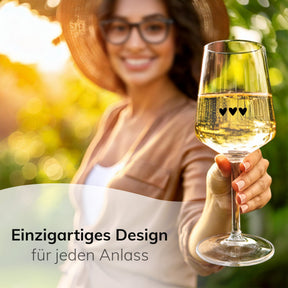 Eine lächelnde Frau mit Sonnenhut und Brille hält ein ElbFuchs Weinglas der Serie 'Midnight Affection' mit drei feinen Herzgravuren in die Kamera, gefüllt mit funkelndem Weißwein, vor sonnigem, grün verschwommenem Gartenhintergrund.