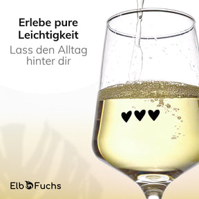 Nahaufnahme eines ElbFuchs Weinglases 'Midnight Affection' mit dezenter Herzgravur, in das gerade spritziger Weißwein eingegossen wird, vor einem hellen, warm getönten Hintergrund.