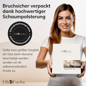 Eine lächelnde Frau in einem weißen ElbFuchs T-Shirt hält die Geschenkbox der Weingläser 'Midnight Affection' in den Händen, daneben das Zufriedenheitsversprechen-Siegel und der Hinweis auf bruchsichere Verpackung durch hochwertige Schaumpolsterung.
