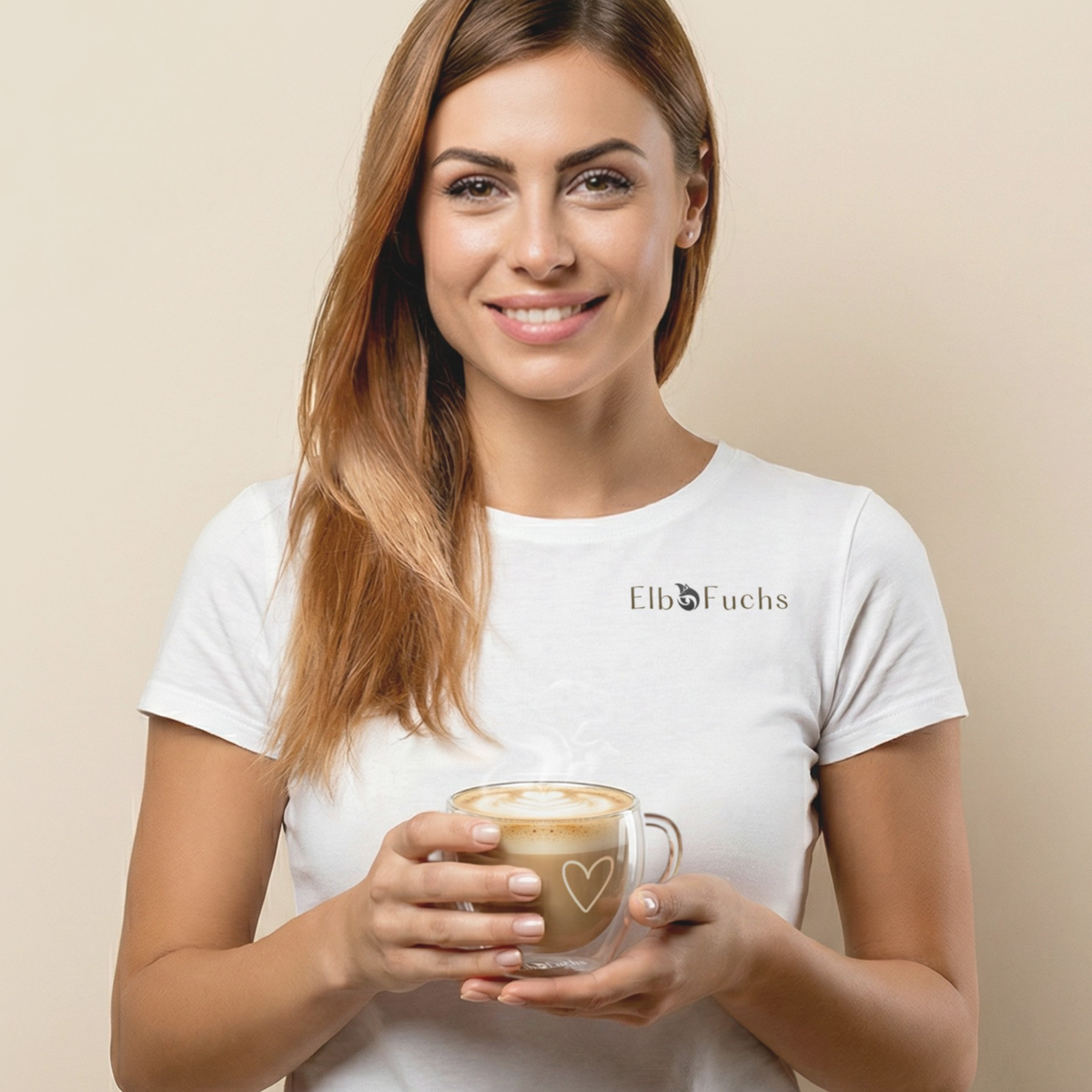 Eine sympathisch lächelnde Frau in einem weißen ElbFuchs T-Shirt hält ein doppelwandiges Kaffeeglas mit Henkel und feiner weißer Herzgravur in beiden Händen. Das Thermoglas ist mit heißem, leicht dampfendem Cappuccino und feiner Latte-Art befüllt. Einladendes Motiv für das ElbFuchs Zufriedenheitsversprechen vor einem hellen, neutralen Studiohintergrund.