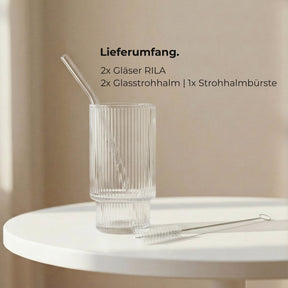 Ein leeres, fein geriffeltes ElbFuchs RILA Glas mit Glasstrohhalm und Reinigungsbürste auf einem weißen Tisch, daneben der Lieferumfang: 2 Gläser, 2 Glasstrohhalme und eine Strohhalmbürste.